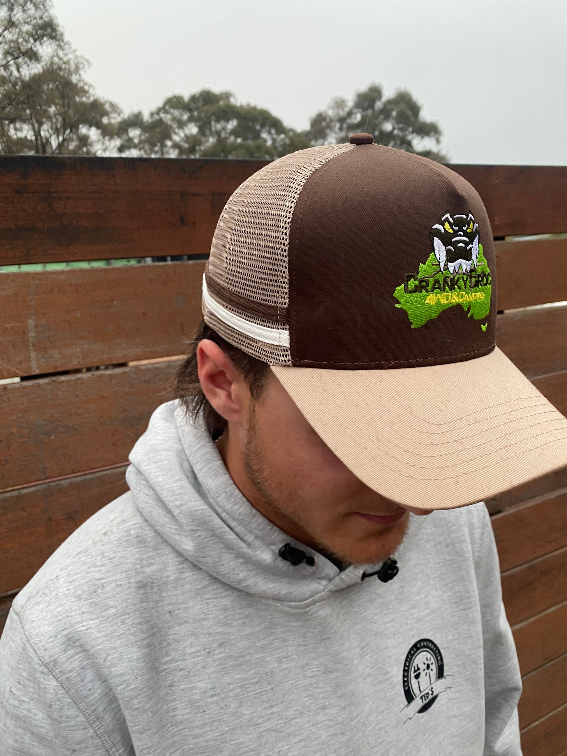 Person wearing a brown trucker hat with a Cranky Croc logo, The trucker cap also has a brown and white stripe on the side with a cream cap. Person is standing against a wooden fence. 
