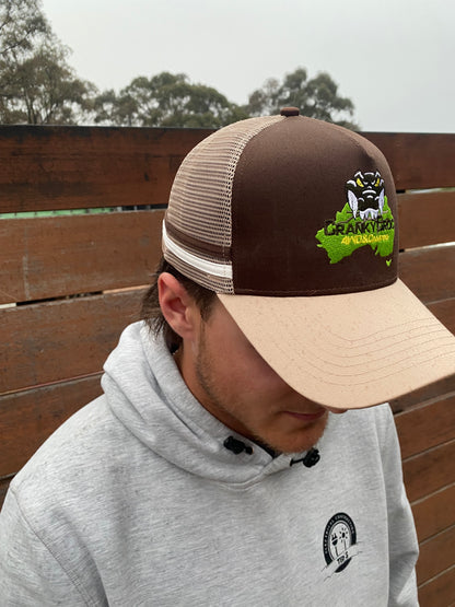 Person wearing a brown trucker hat with a Cranky Croc logo, The trucker cap also has a brown and white stripe on the side with a cream cap. Person is standing against a wooden fence. 