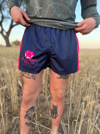 Front view of person wearing navy and pink Aussie Footy Shorts with zip pockets with a Cranky Croc logo and crocodile design. Photo is set in a field