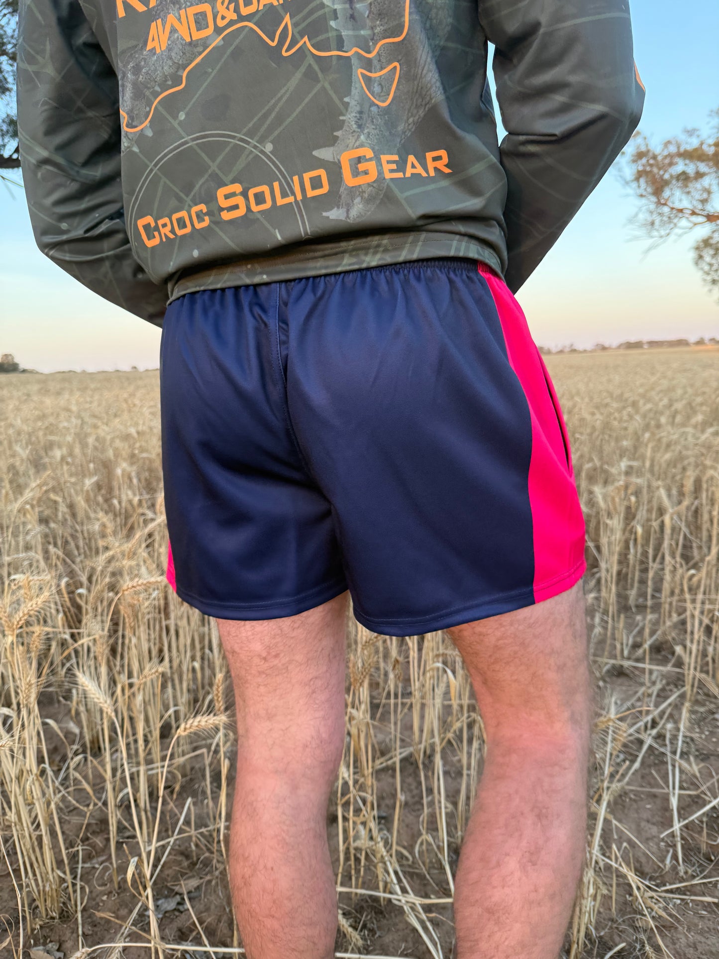 Back view of person wearing navy and pink Aussie Footy Shorts with zip pockets with a Cranky Croc logo and crocodile design. Photo is set in a field