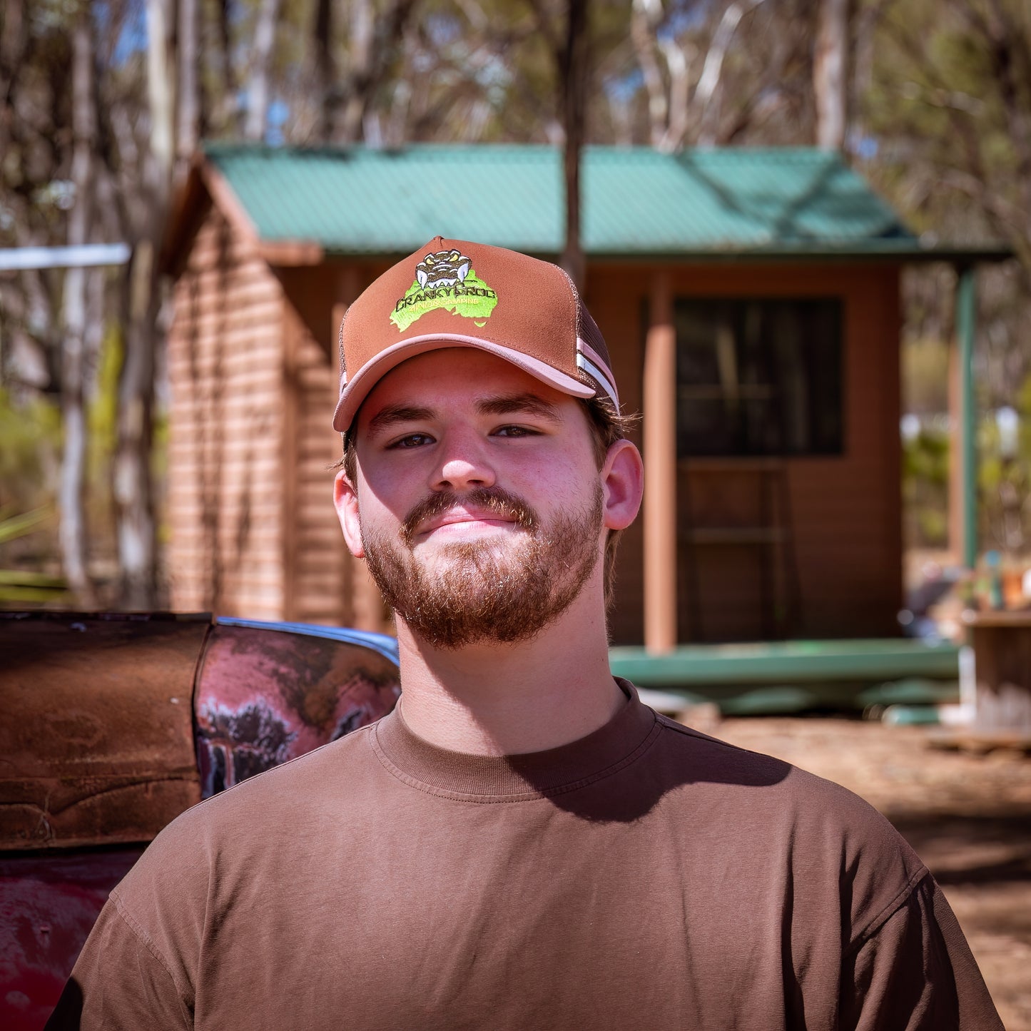 Man wearing a pink with brown trucker cap and shirt outdoors with a wooden cabin in the background