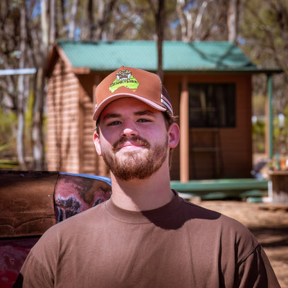 Man wearing a pink with brown trucker cap and shirt outdoors with a wooden cabin in the background