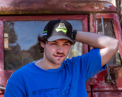 Man wearing a black trucker cap in front of an old red truck. The country trucker cap also has a green and white stripe across the sides and a crocodile design on the front