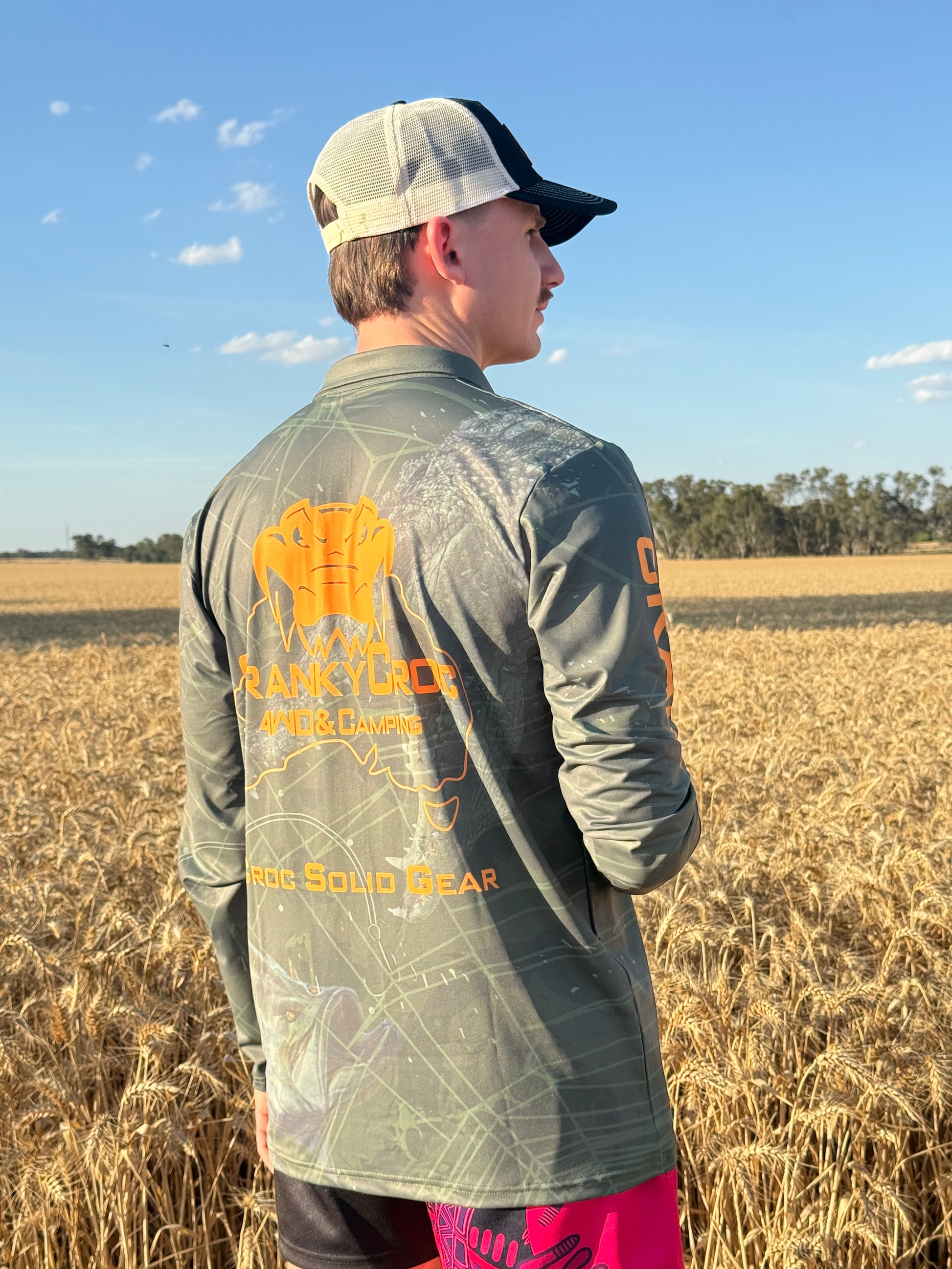 Man wearing dark green long sleeve fishing shirt / hunting shirt. The fish shirt has a fluro orange 'Cranky Croc' logo on the back with a crocodile and barramundi design. Man wearing the camo fishing shirt is standing in a crop field