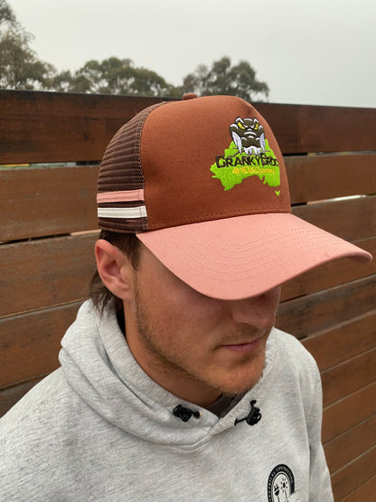 Man wearing a light pink and brown trucker cap next to a timber fence. The cap also has a pink and white stripe on the side.