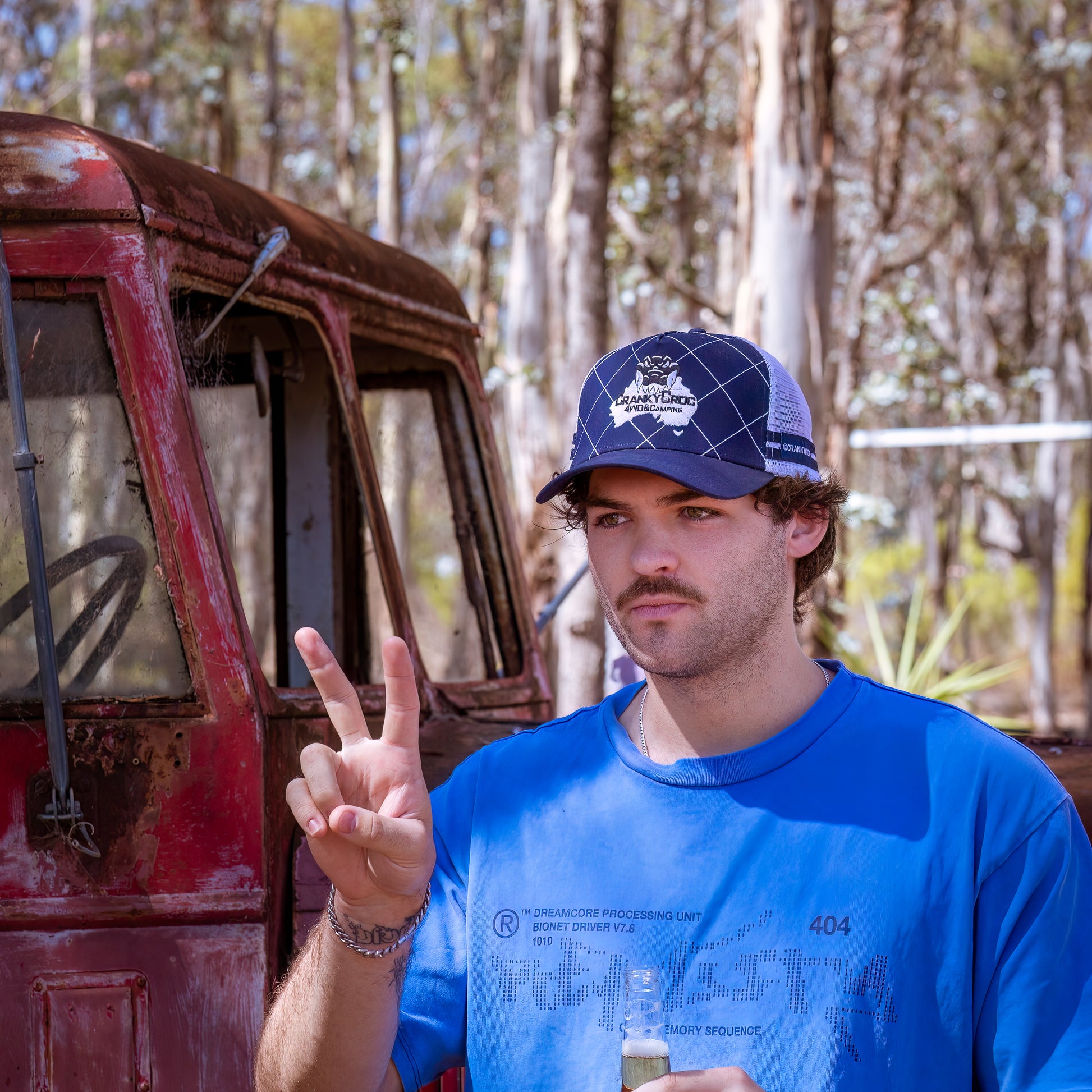 Man wearing a Navy trucker cap making a peace sign in front of a rusted truck.