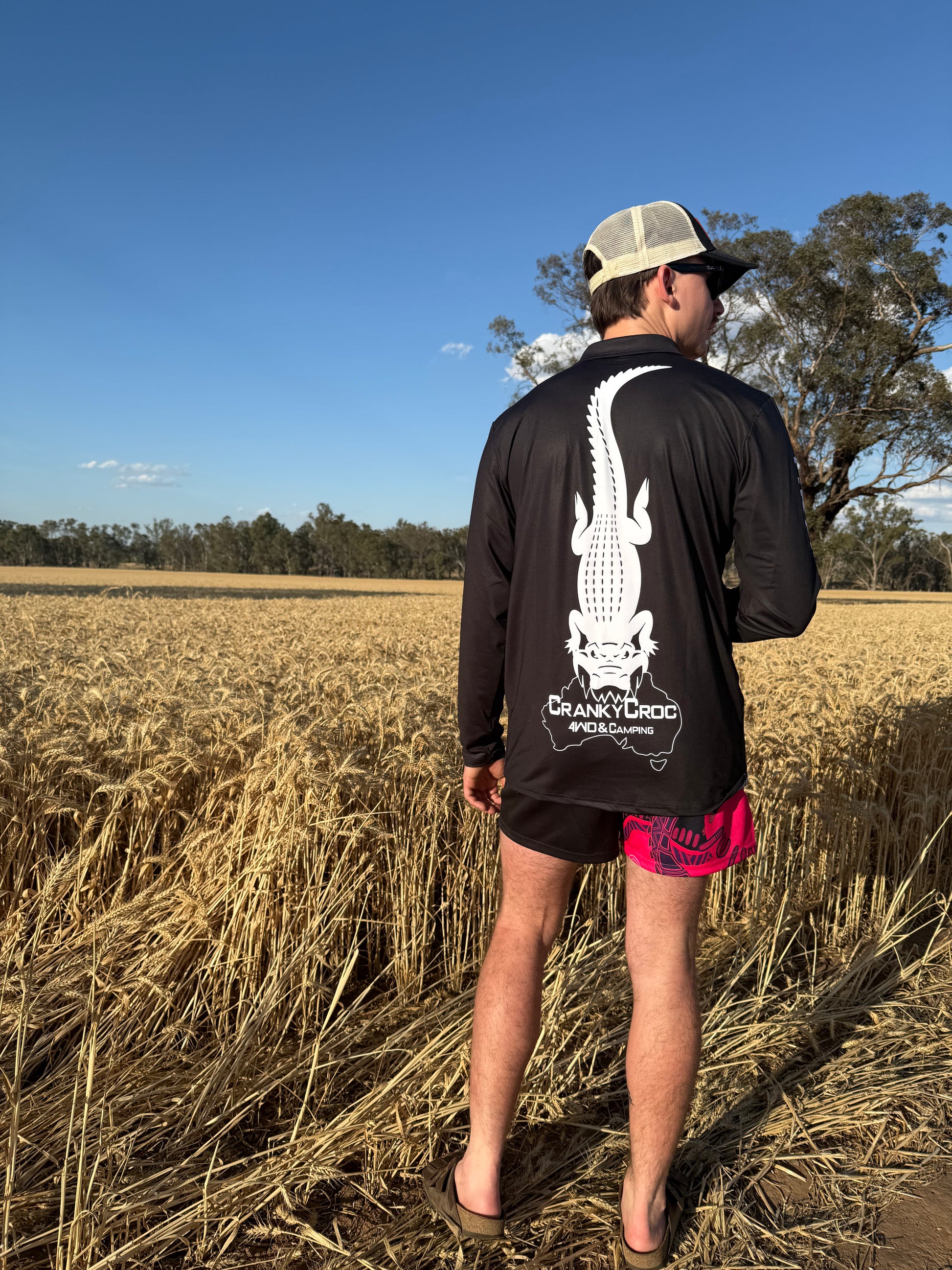 Man with his back facing wearing a black long sleeve fishing shirt with a white crocodile design and logo. man is standing in a crop field.