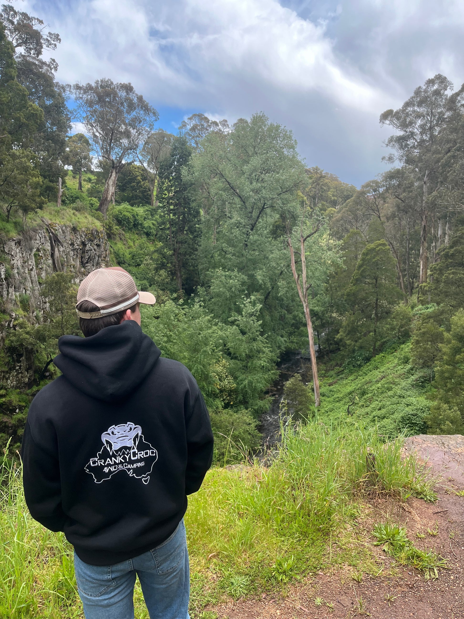 Person wearing a black hoodie and with a white embroidered logo, and wearing a trucker cap. Man is standing on a cliff in bush land area.