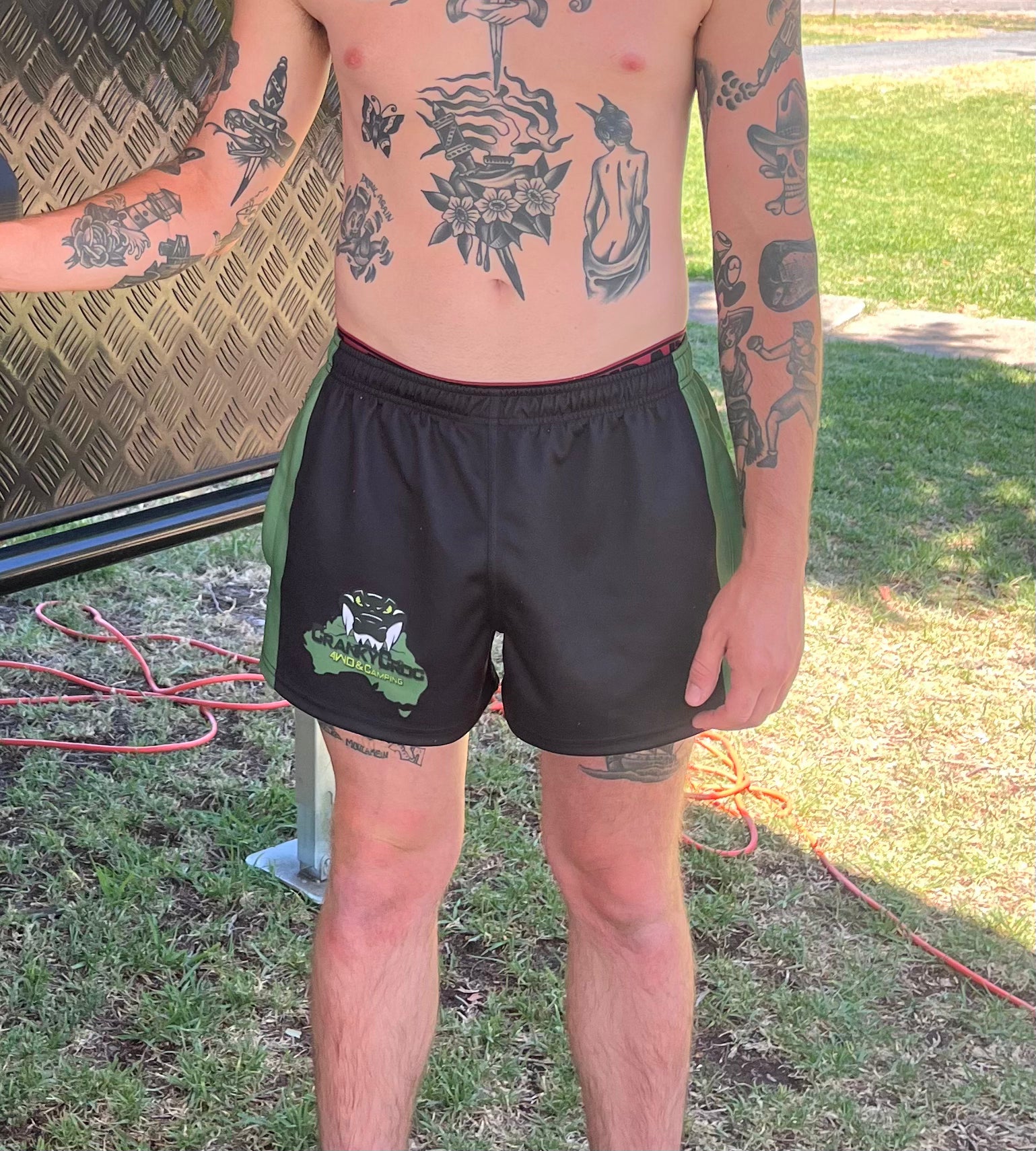 Man wearing black with green Aussie Footy Shorts with a logo, standing outdoors on grass.