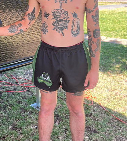 Man wearing black with green Aussie Footy Shorts with a logo, standing outdoors on grass.