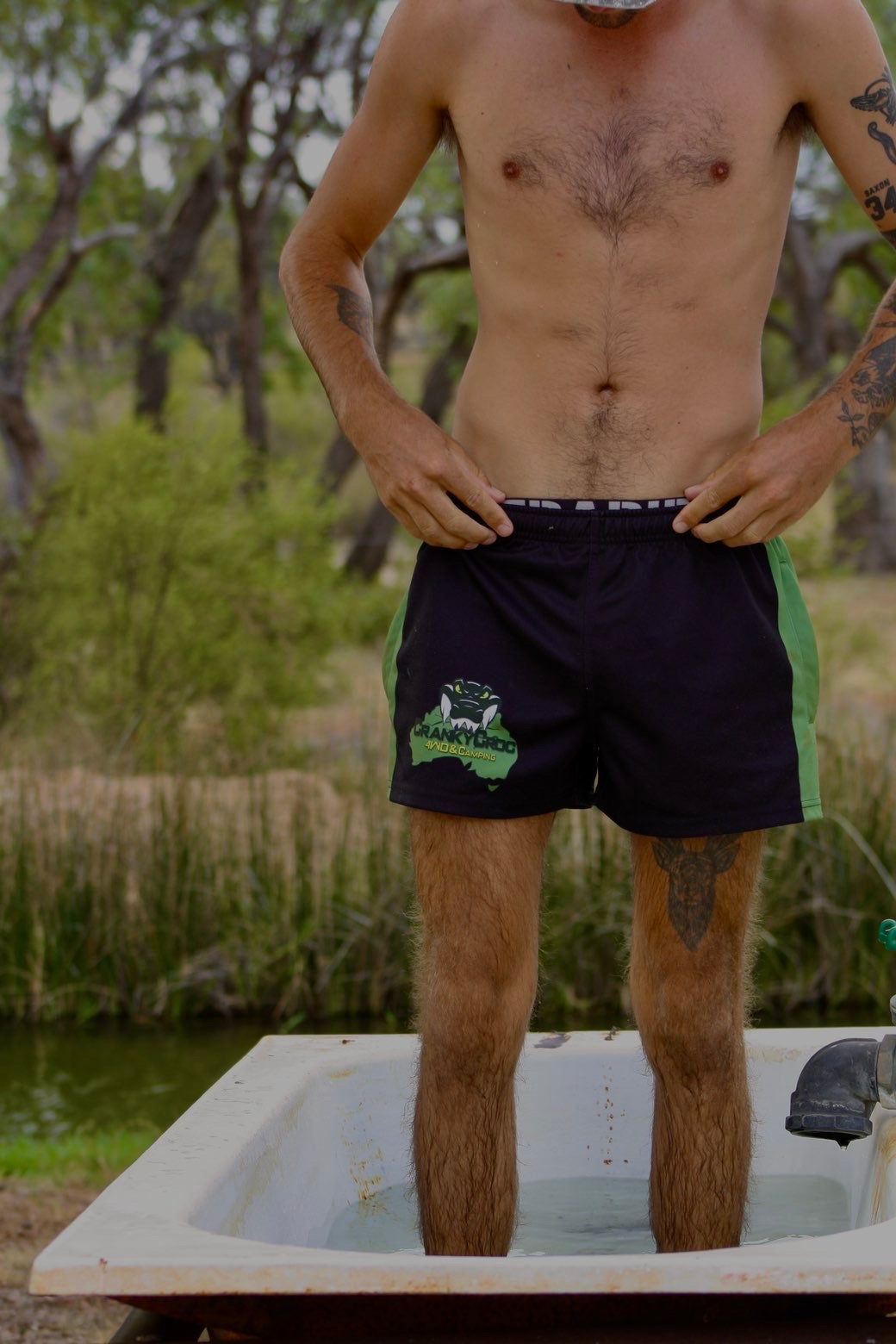Man standing in a bathtub outdoors with a natural background wearing green with black Aussie Footy Shorts