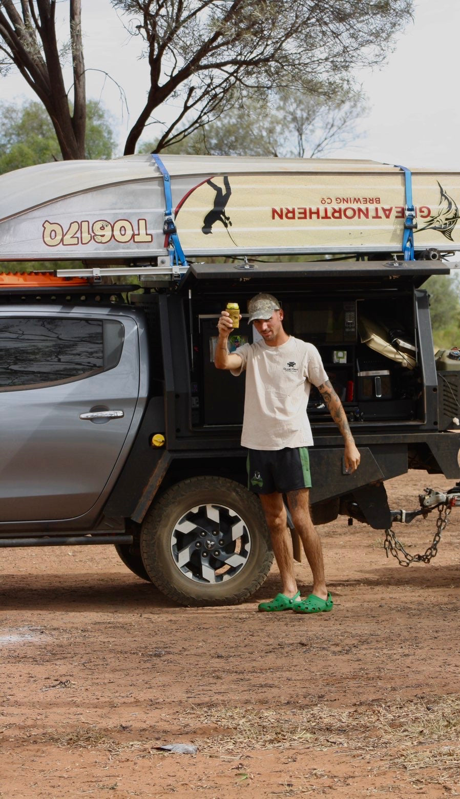Man wearing black with green Aussie Footy Shorts standing next to a 4WD holding a beer
