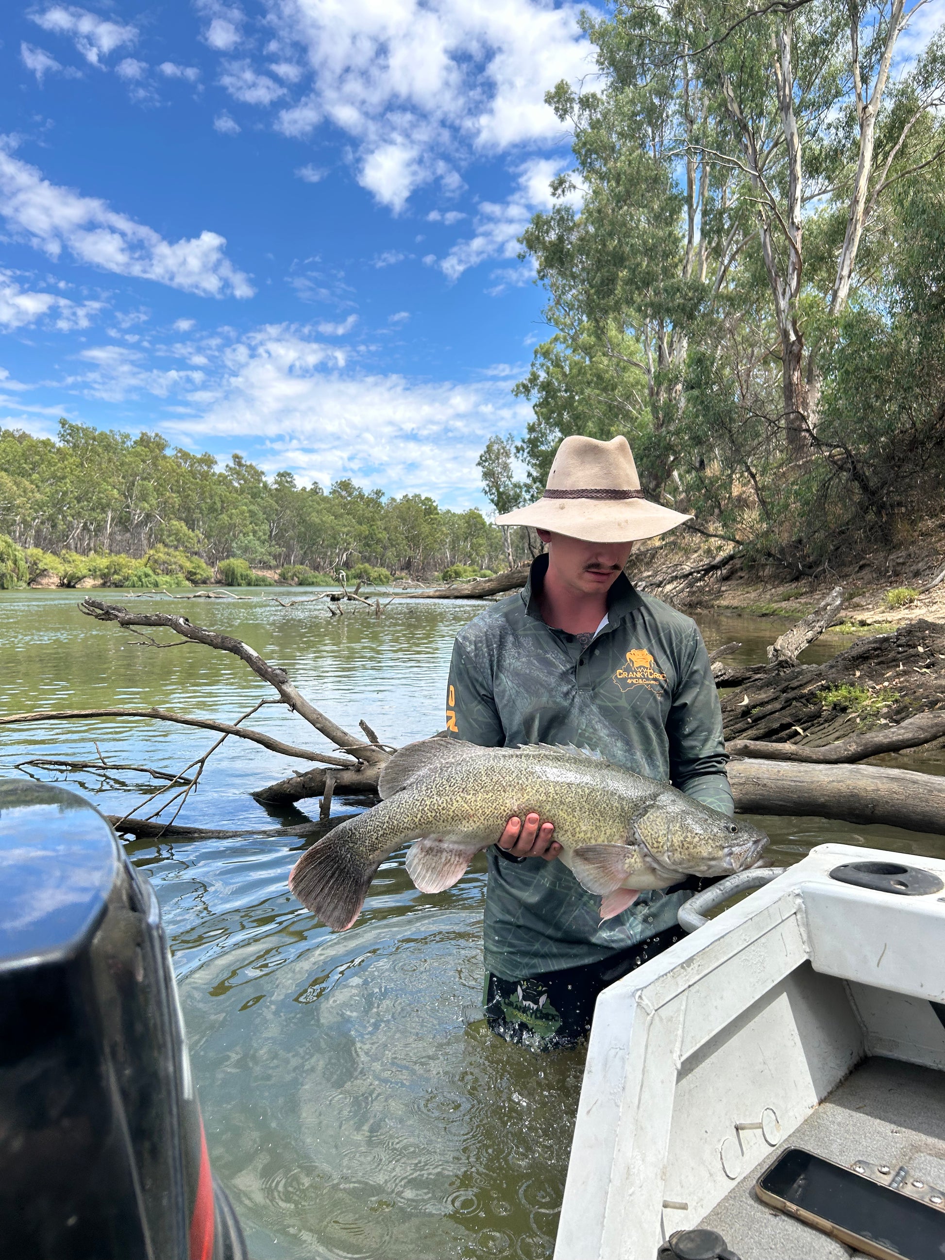 Man wearing dark green long sleeve fishing shirt / hunting shirt while holding a large Murray Cod fish. The fish shirt has a fluro orange 'Cranky Croc' logo on the front with a crocodile and barramundi design. Man wearing the camo fishing shirt is standing in a river next to a fishing boat and is wearing a Akubra hat