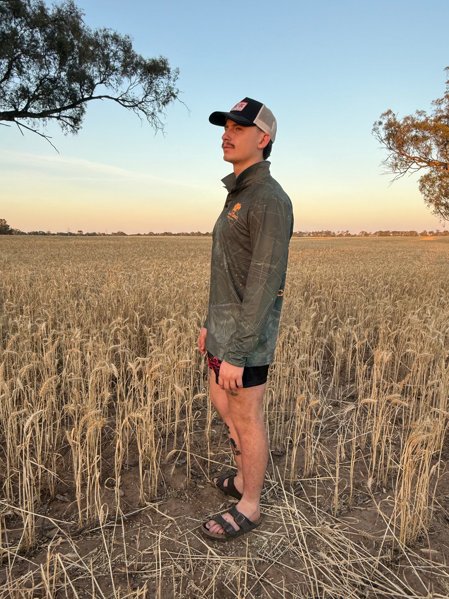 Man wearing a dark green long sleeve fishing shirt / hunting shirt. The fish shirt has fluro orange 'Cranky Croc' branding with a crocodile and barra design standing a field at sunset 