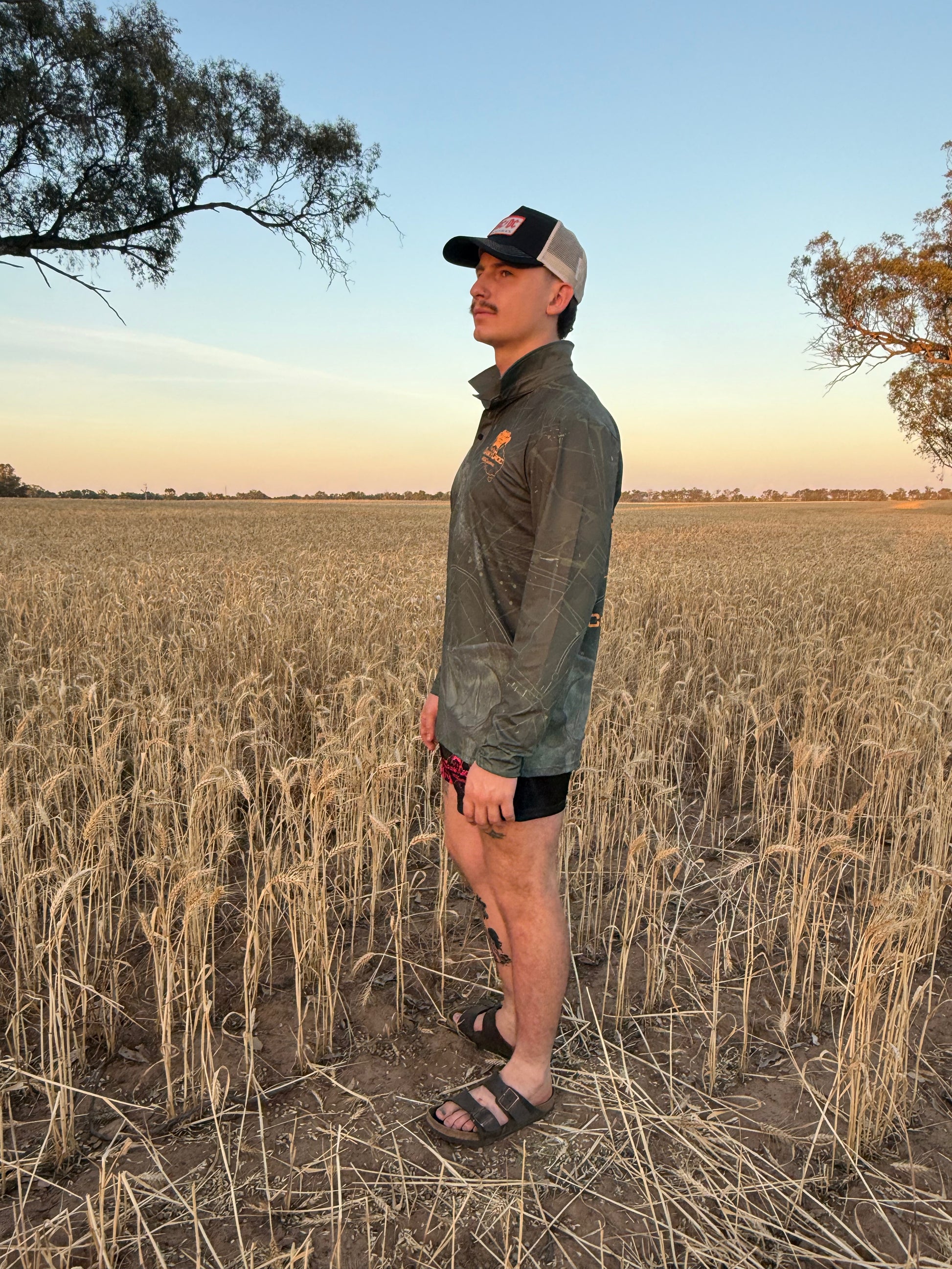 Man wearing a dark green long sleeve fishing shirt / hunting shirt. The fish shirt has fluro orange 'Cranky Croc' branding with a crocodile and barra design standing a field at sunset 