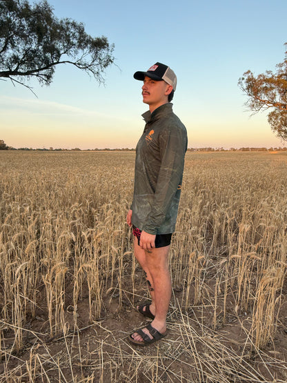 Man wearing a dark green long sleeve fishing shirt / hunting shirt. The fish shirt has fluro orange 'Cranky Croc' branding with a crocodile and barra design standing a field at sunset 