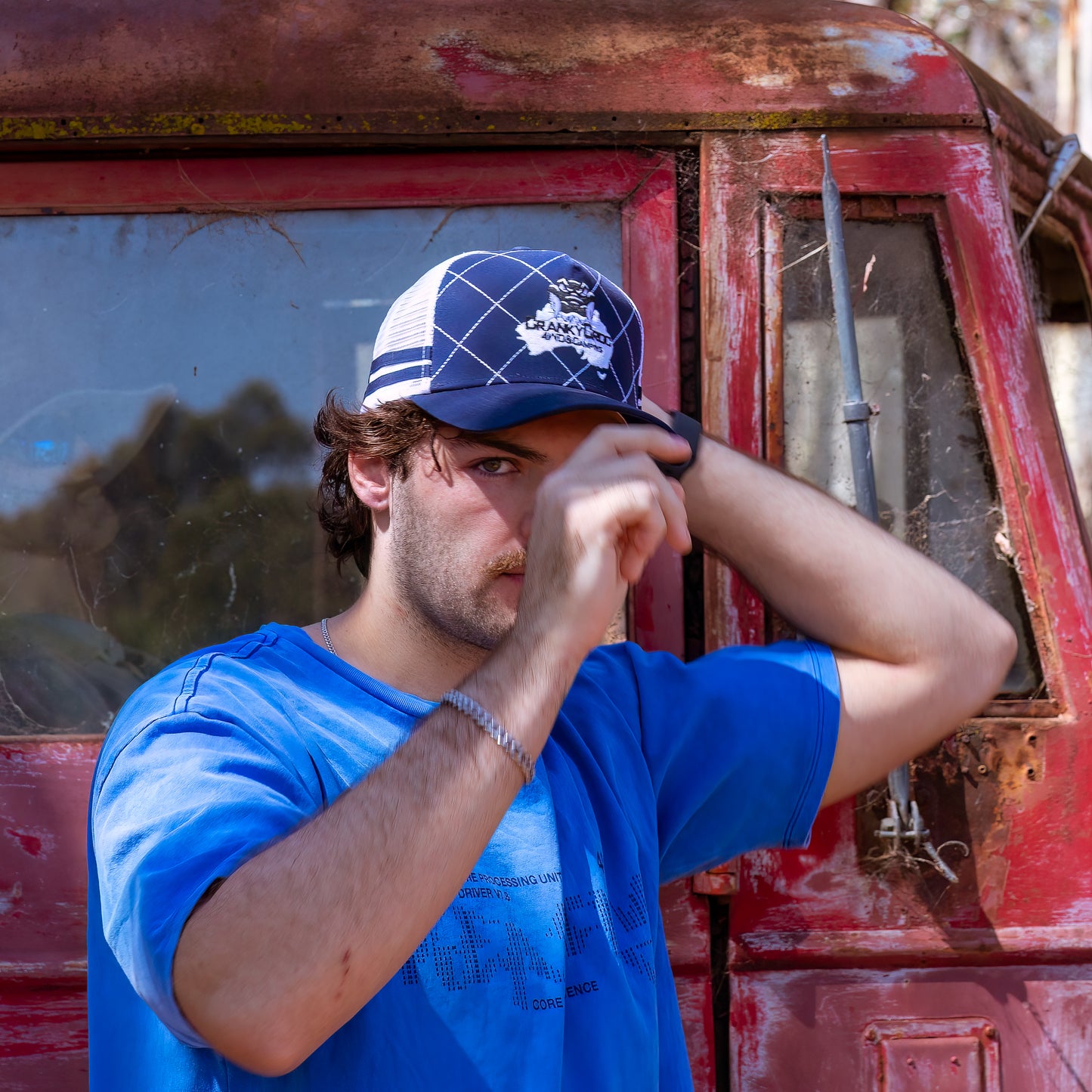 Man standing infront of a red truck wearing a navy Country Trucker cap with a logo. The Trucker Hat is coloured navy with white.