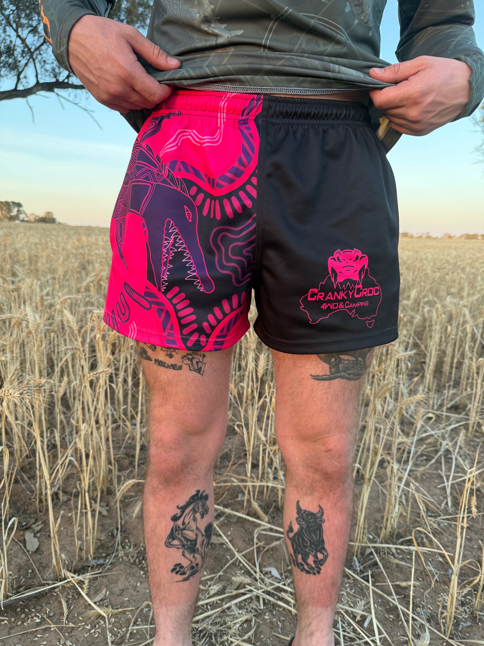 Front view of person wearing pink with black indigenous footy shorts with zip pockets with a Cranky Croc logo and crocodile indigenous art design. Photo is set in a field