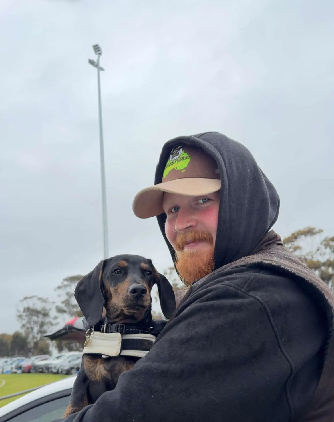 Man with a dachshund dog on a cloudy day wearing a brown trucker hat with a cranky croc logo on the front.