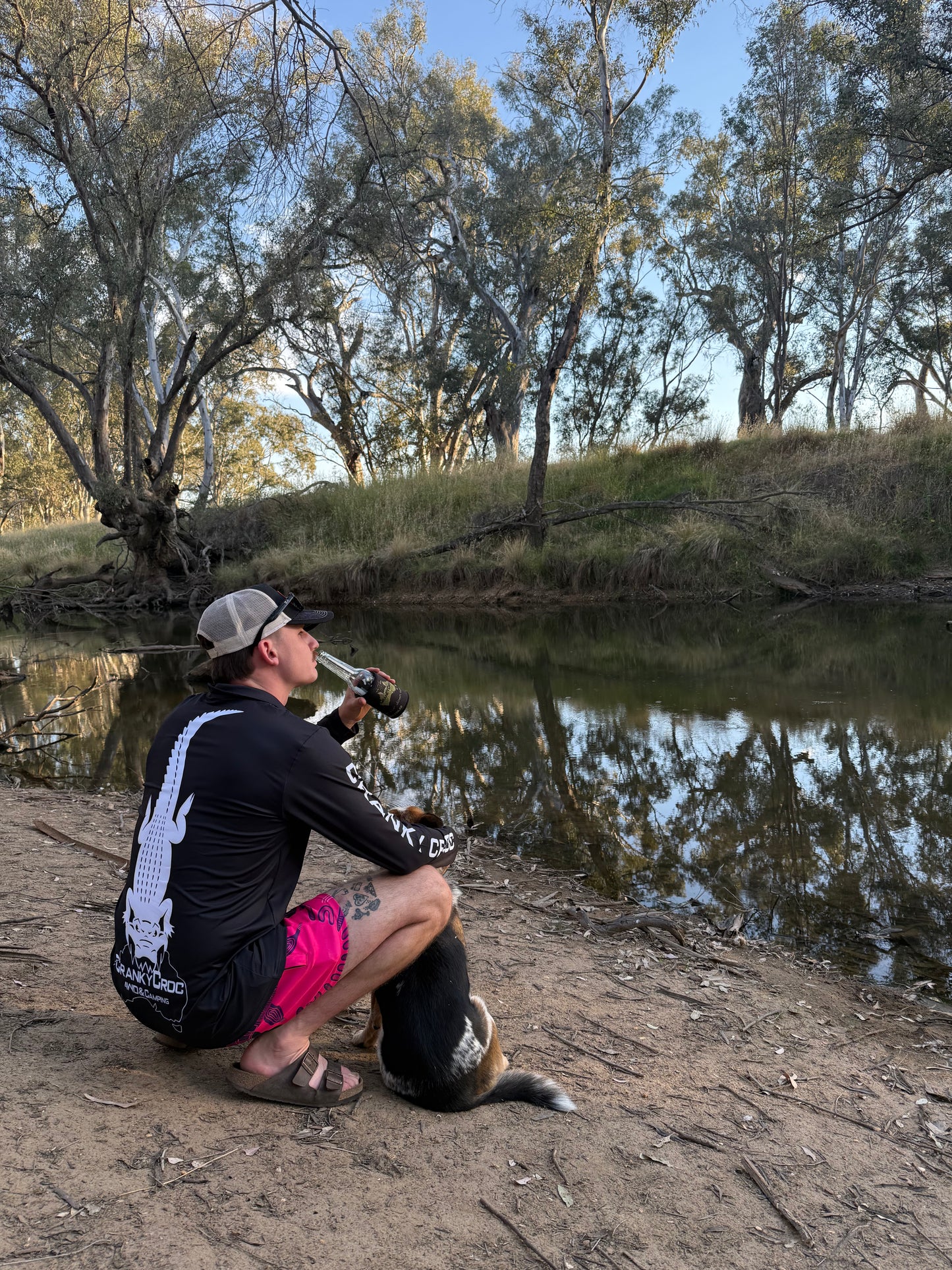 Man with a dog by a river with trees in the background wearing Aussie footy shorts and a black long sleeve fishing shirt with a white crocodile design.
