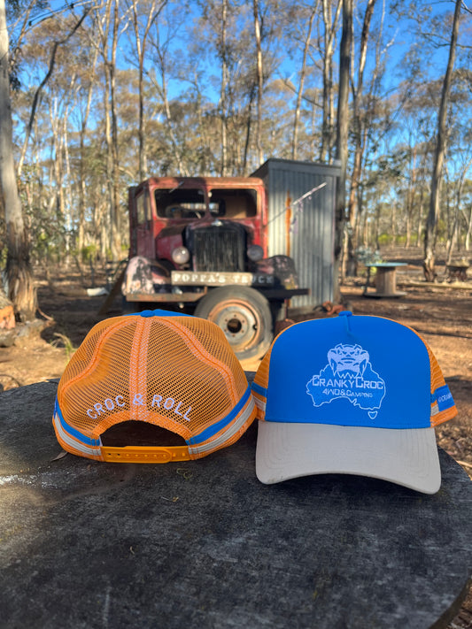 Two country trucker hats showing off the front and back. The country trucker caps are orange with blue with a crocodile design, on a rock with a vintage truck and shed in the background.