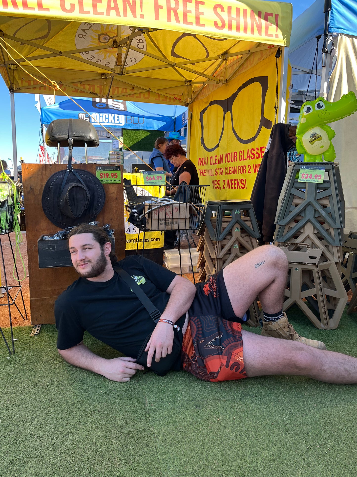 Person wearing Aussie Footy Shorts lying on the ground in front of a promotional stand with cleaning products for glasses.