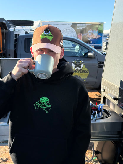 Person wearing a black hoodie and brown trucker cap with a Cranky Croc logo, holding a white mug outdoors.