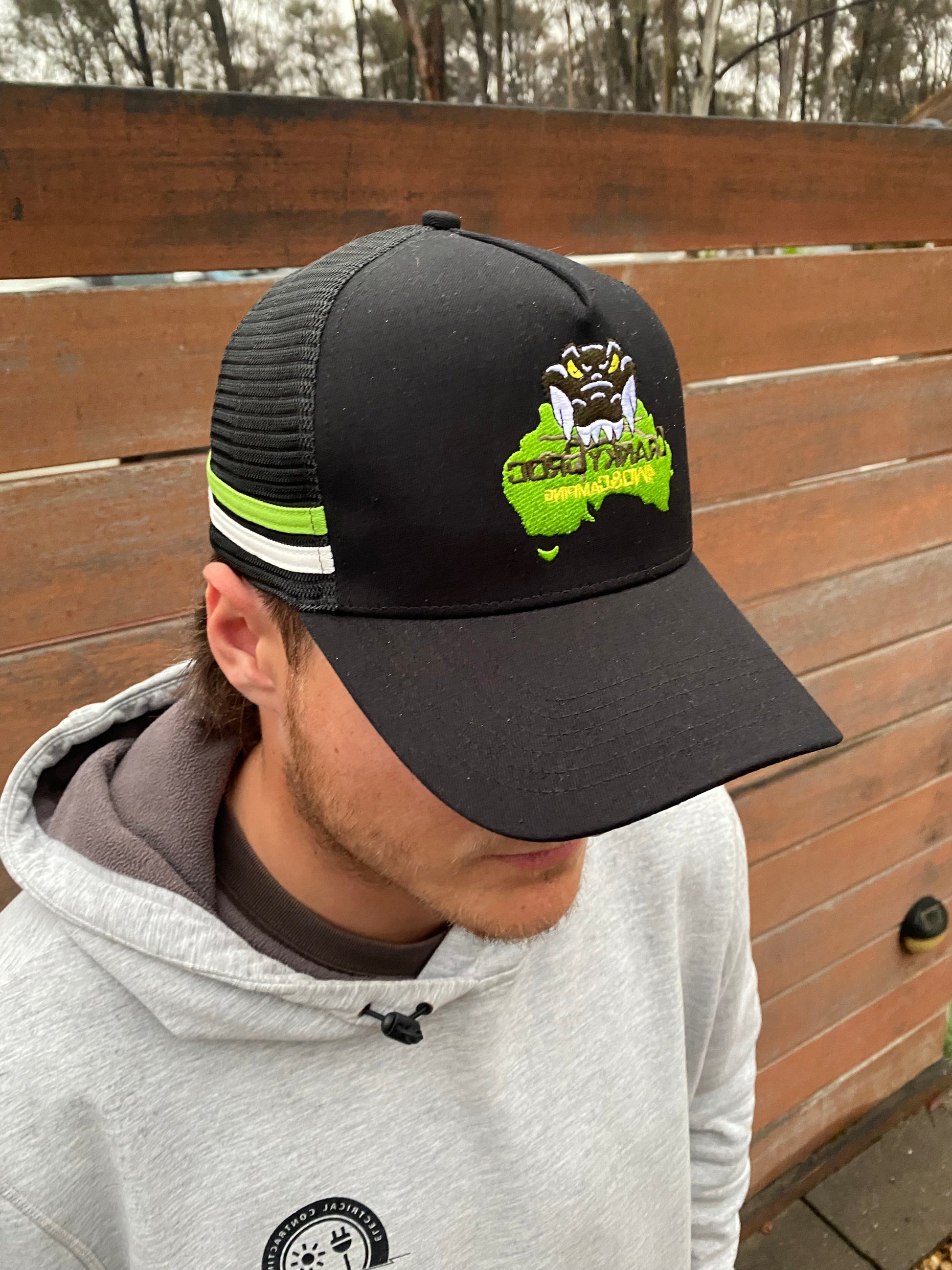 Person wearing a black trucker hat with a Cranky Croc logo, The trucker cap also has a green and white stripe on the side. Person is standing against a wooden fence. 