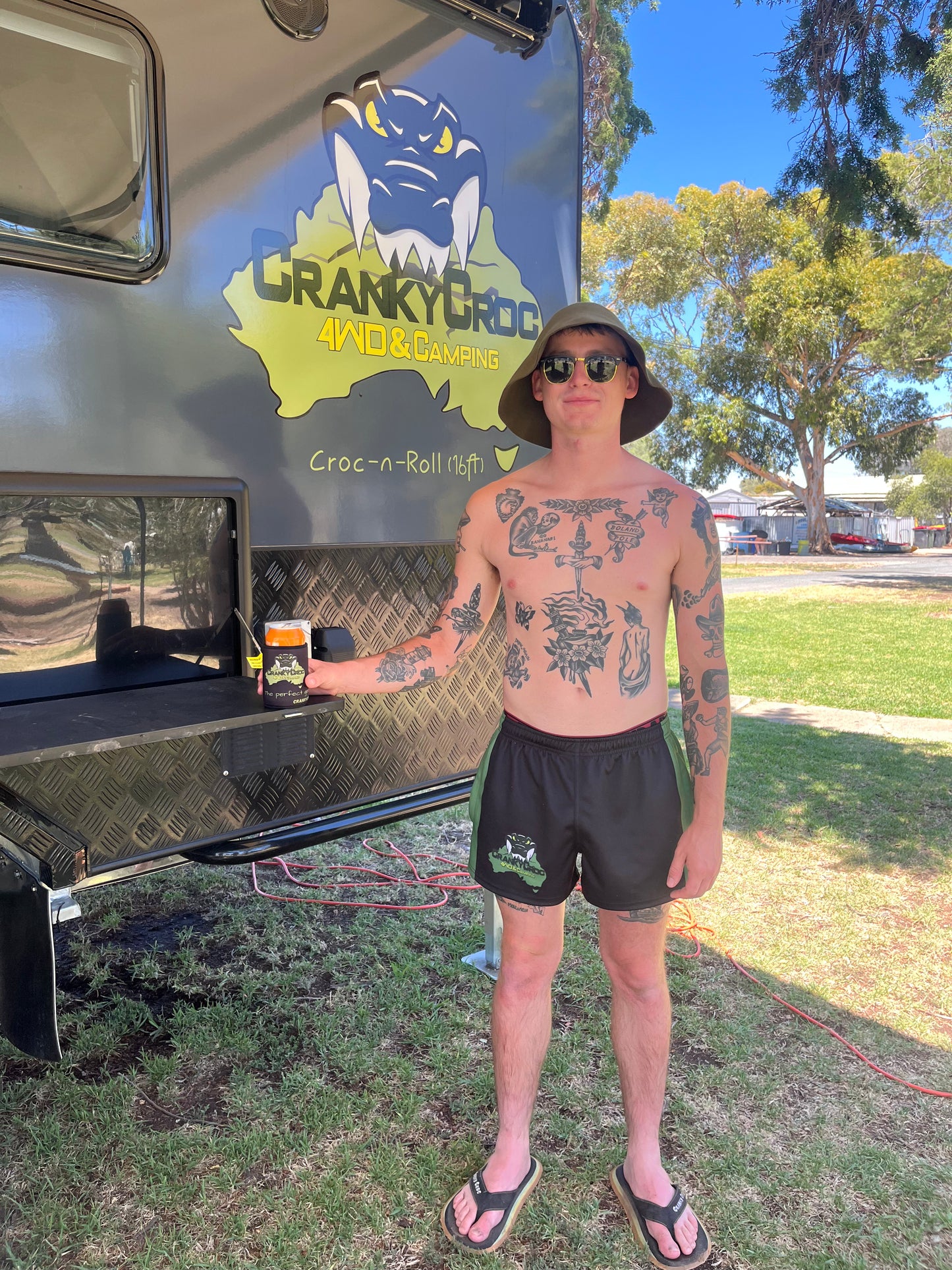 Man with tattoos wearing black with green Aussie Footy Shorts standing next to a vehicle with 'Cranky Croc' branding. He is wearing a pair of Cranky Croc Great Aussie Pluggers – top of pluggers are black and coloured on the sides.
