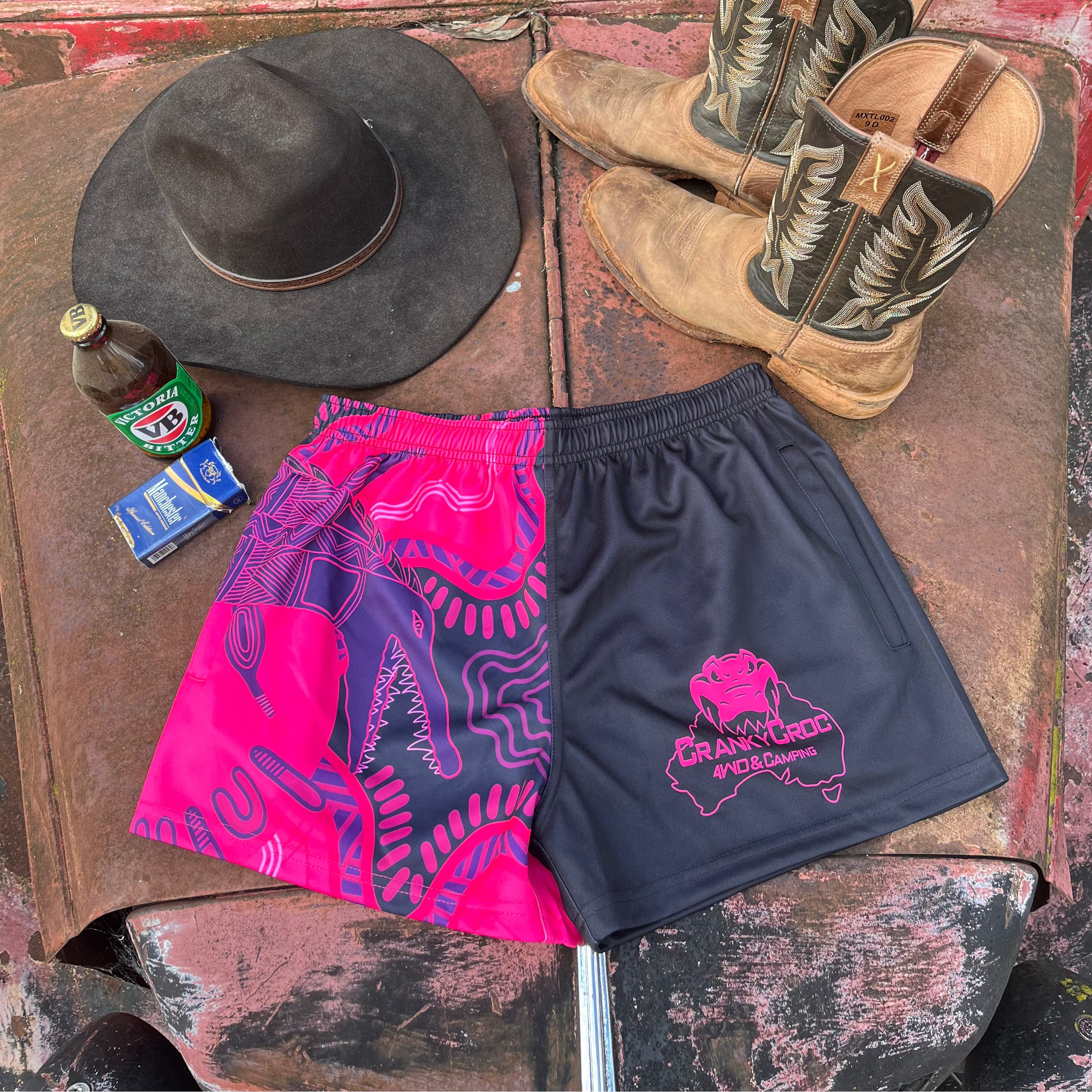 Pink Indigenous Country Aussie Footy Shorts on a 4wd bonnet next to a pair of country boots, country cowboy hat and a beer.