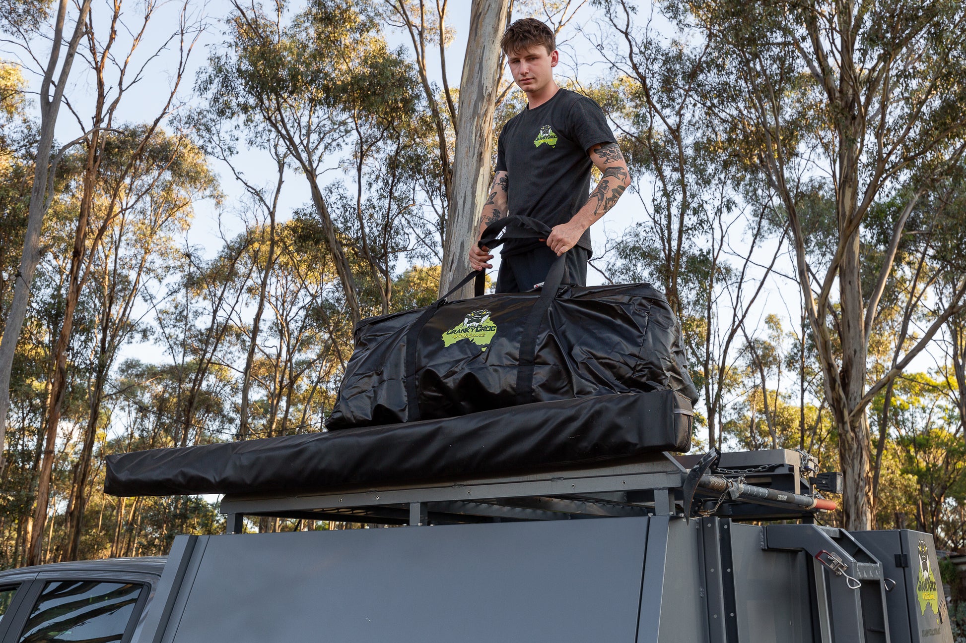 A person standing on a vehicle with a large black PVC camping bag loaded on top.