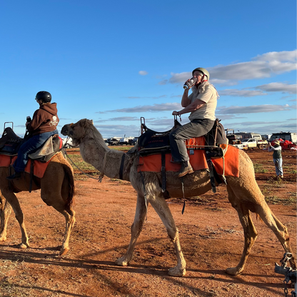 A woman riding a camel drinking a beer out of her TWO UP two can can holder