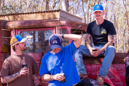 Three men sitting on a red truck, wearing blue trucker caps and also navy trucker hats and casual clothing, while holding a beer.