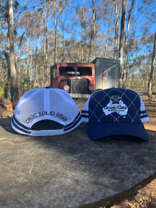 Two Country Trucker Caps with visible branding showing both front and back of hats on a wooden surface with a rustic truck and trees in the background. The trucker hats are coloured navy with white.