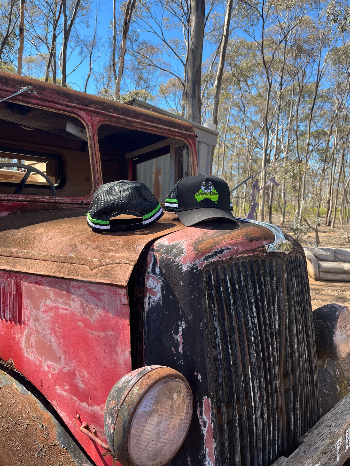 Two black trucker caps on an old, rusted truck in a forest setting. The country trucker caps also have a green and white stripe down the sides