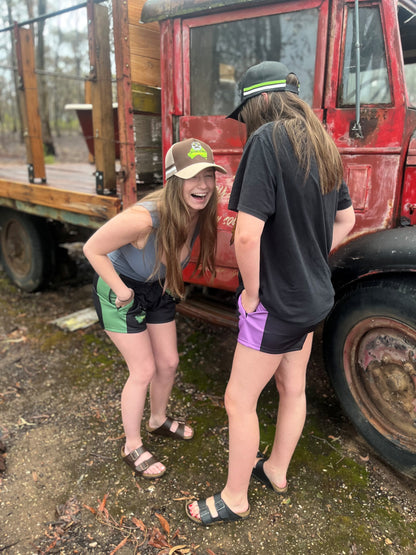 Two girls laughing while wearing Aussie Footy Shorts. One of the girls is wearing a brown trucker hat and the other is wearing a black trucker hat. They are standing next to an old truck.