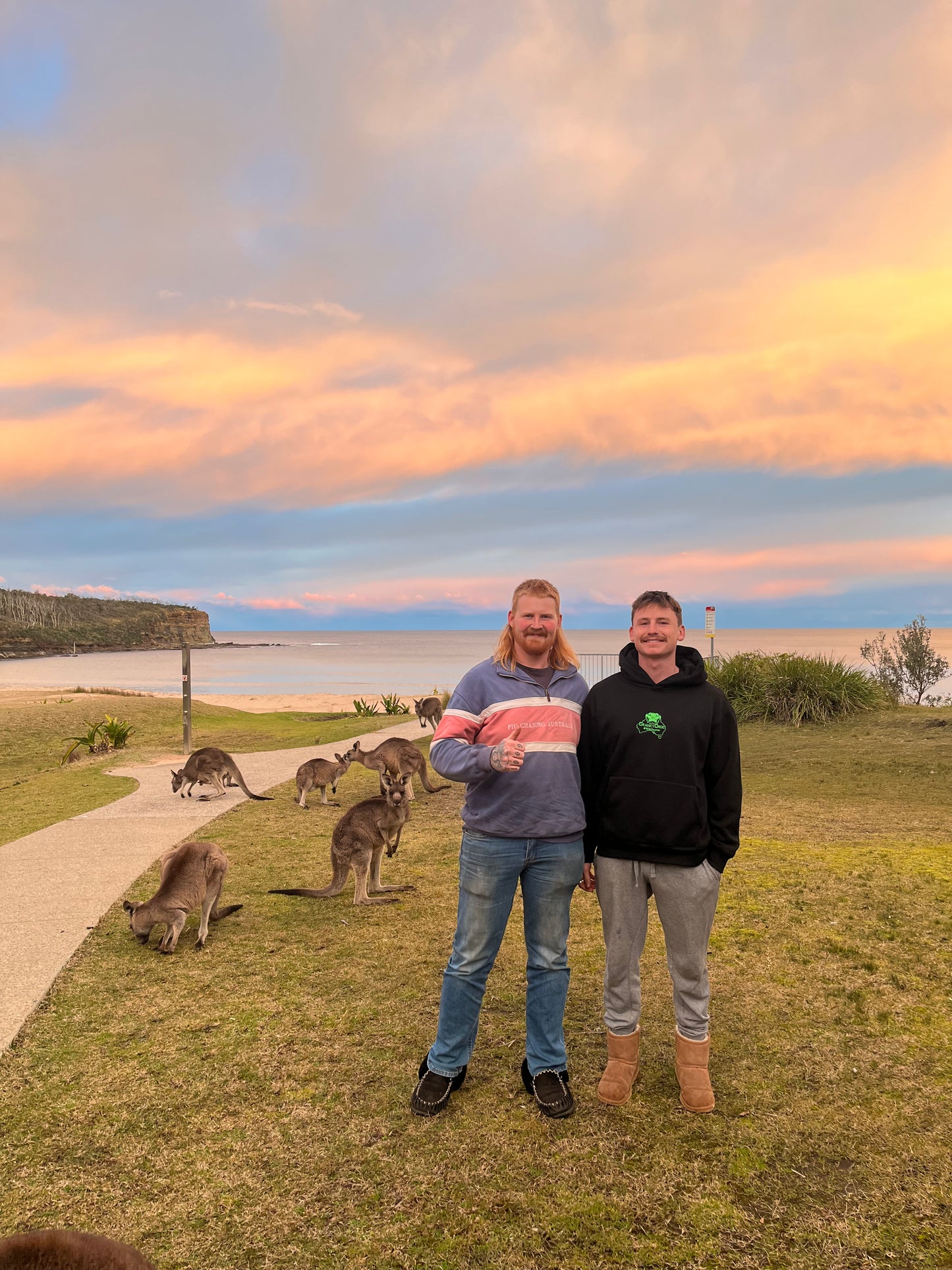Two people standing on a grassy area with kangaroos near a beach at sunset. wearing a black hoodie by Cranky Croc