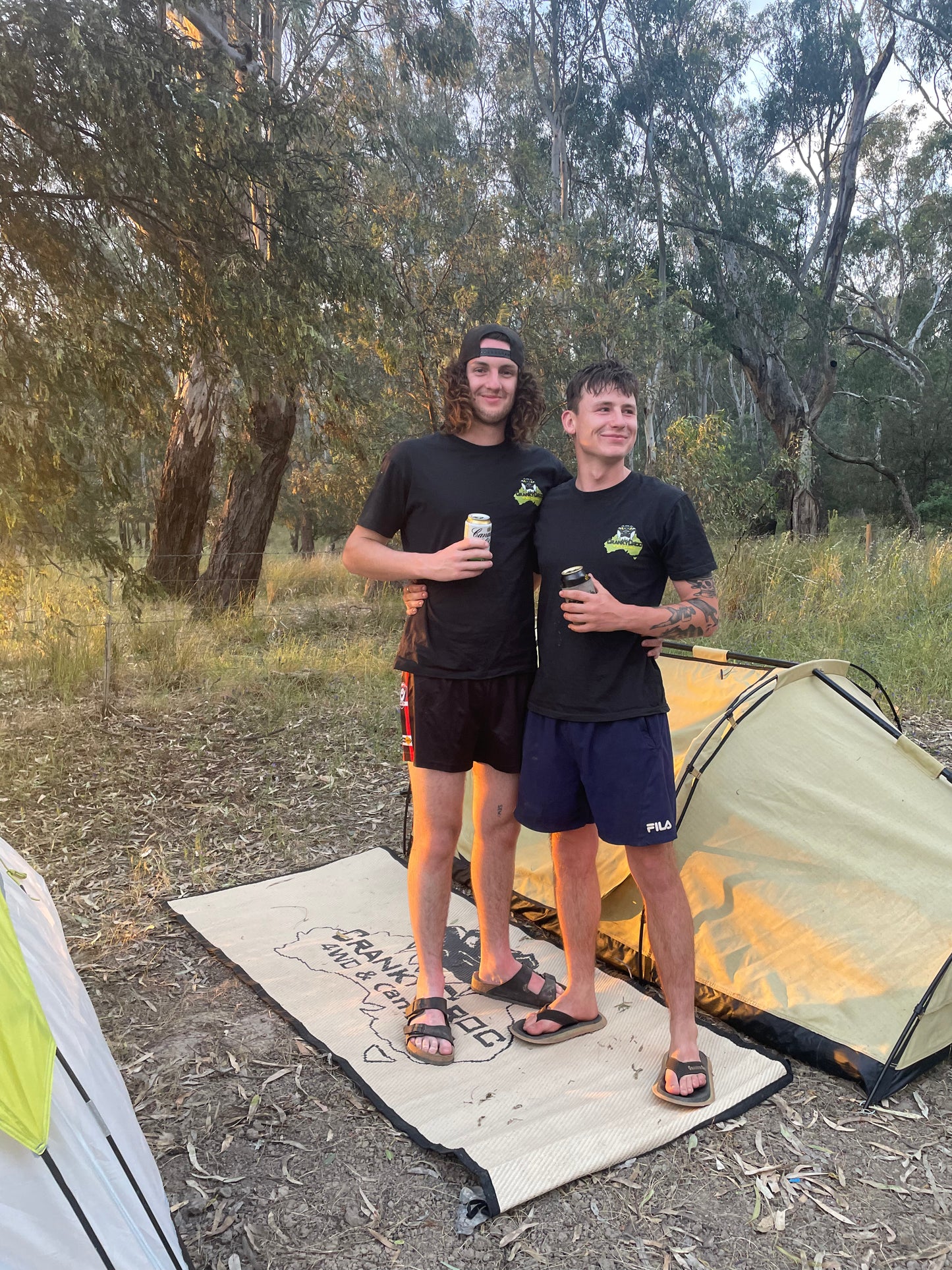 Two people standing outdoors next to a swag in a camping setting. the two men are wearing black tees branded Cranky Croc