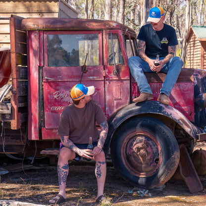 Two men sitting on an old rusted truck wearing Country trucker hats. Both are orange with blue trucker caps