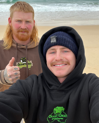 Two men on a beach with ocean waves in the background. The men are wearing black embroidered hoodies 