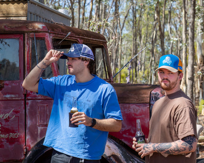 Two men standing in front of an old red truck with trees in the background wearing a white with navy trucker cap and a orange with blue trucker cap