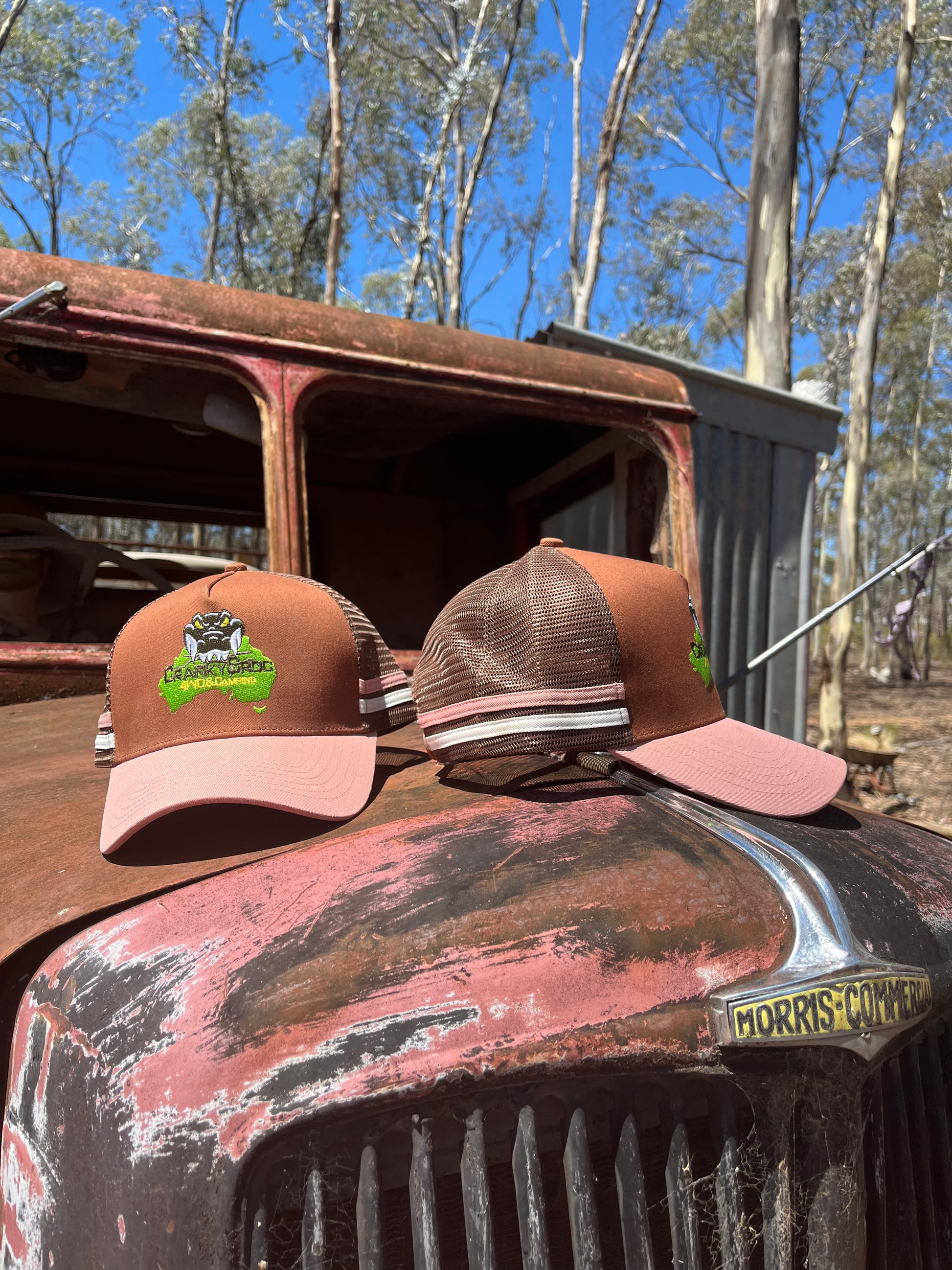 Two Country Trucker Caps on a rusted vintage truck with trees in the background. Both are brown with pink trucker hats
