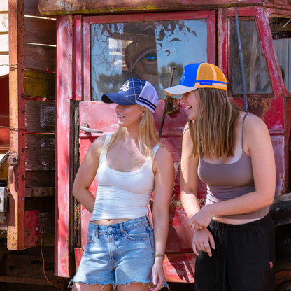 Two women standing in front of a red truck, wearing Country Trucker hats. One is wearing a light blue with orange trucker cap, the other is wearing a white with navy trucker cap