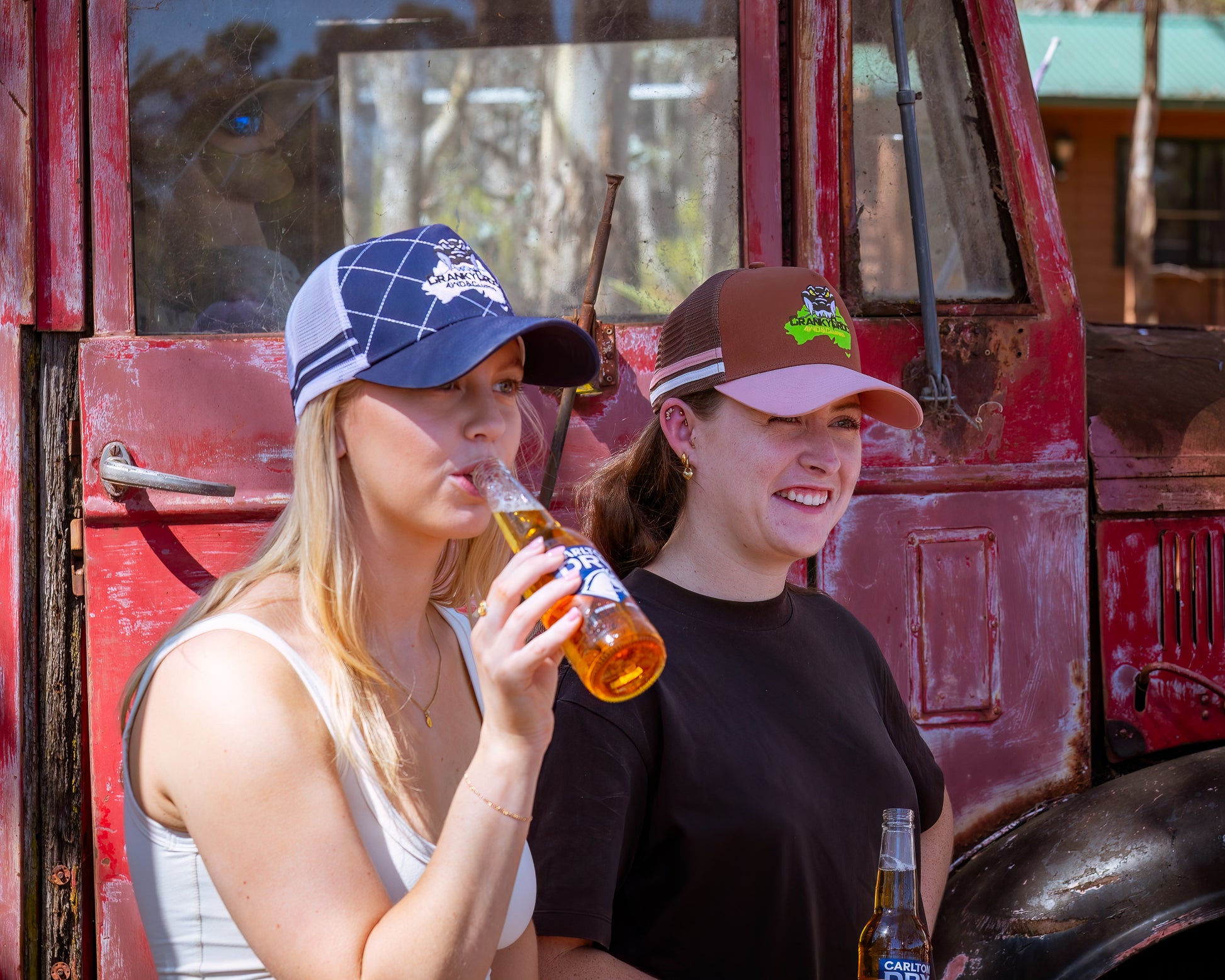 Two women standing in front of a red truck, one drinking from a beer bottle. One girl is wearing a white with navy trucker hat, and the other is wearing a pink with brown trucker hat.