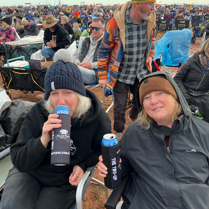 Two woman sitting on Cranky Croc camp chairs at a concert drinking beer from their two can, stubby holders