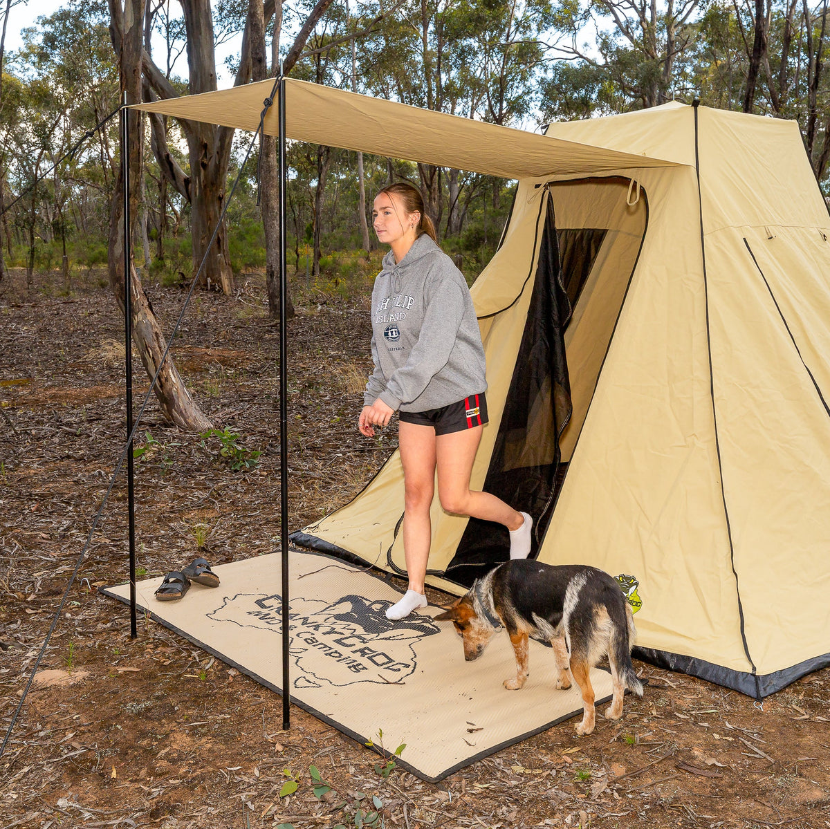 A woman walks out of a beige camping safari tent with a matching swag mat and dog in a wooded outdoor setting.