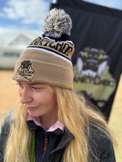 Person wearing a beige warm beanie with a pom pom and a cranky croc logo, standing in front of a black banner.