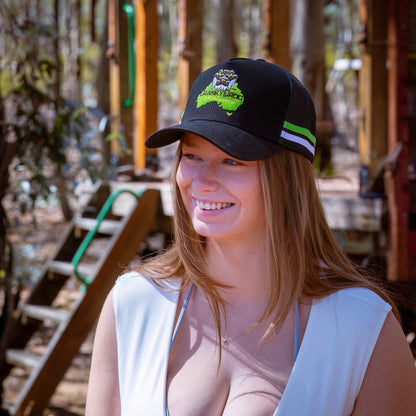 Woman wearing a black trucker cap with a logo, standing outdoors with wooden structures and plants in the background. The Country trucker cap has a green and white stripe across the sides with a Crocodile design on the front