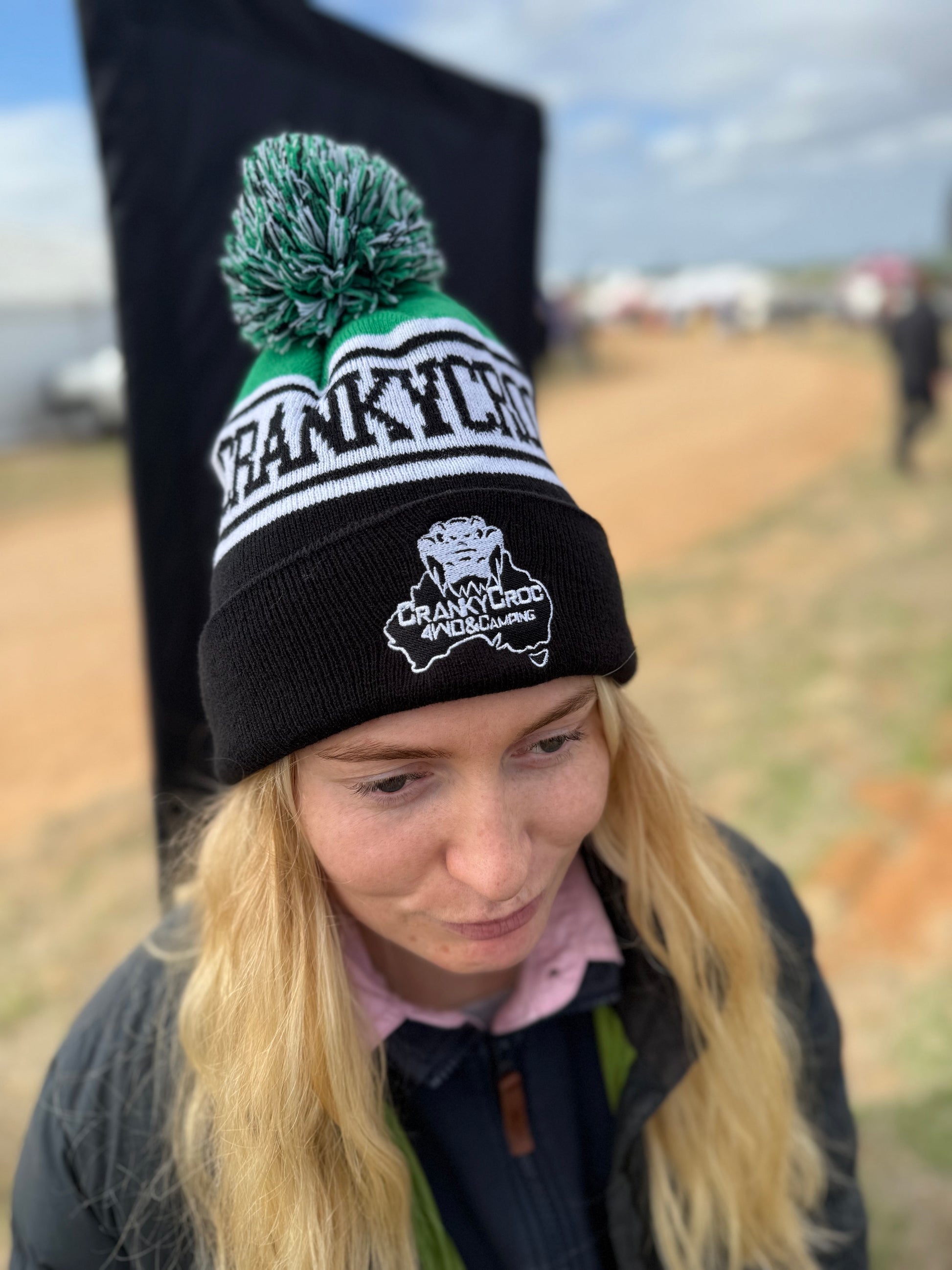 Person wearing a pom pom beanie. The black with green beanie has a 'Crankycroc' branding in an outdoor setting.