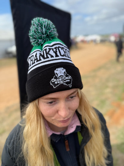 Person wearing a pom pom beanie. The black with green beanie has a 'Crankycroc' branding in an outdoor setting.
